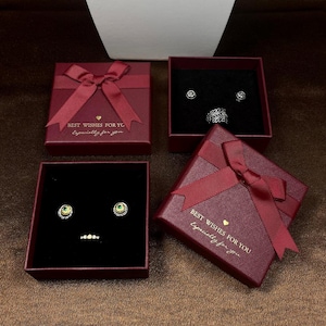 May include: Four burgundy gift boxes with red ribbon bows. Two boxes are open, displaying jewelry on a black velvet interior. The text "BEST WISHES FOR YOU Especially for you" is printed on two boxes. The jewelry includes earrings and a ring.