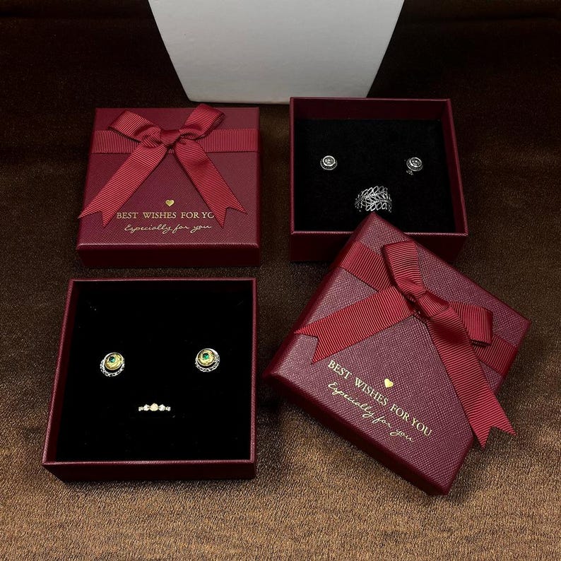 May include: Four burgundy gift boxes with red ribbon bows. Two boxes are open, displaying jewelry on a black velvet interior. The text "BEST WISHES FOR YOU Especially for you" is printed on two boxes. The jewelry includes earrings and a ring.