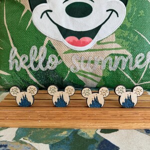 May include: Four wooden Mickey Mouse head ornaments with a blue and white silhouette of Cinderella's castle and fireworks. The ornaments are on a wooden surface.