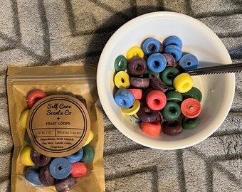 May include: A bag of colorful wax melts shaped like fruit loops. The bag is labeled "Self Care Scents Co. Fruit Loops". The wax melts are in a white bowl on a gray fuzzy surface.