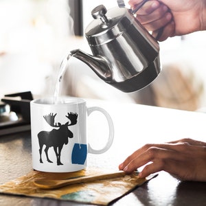 May include: A white ceramic mug with a black silhouette of a moose and a blue tea bag. A silver teapot is pouring hot water into the mug.