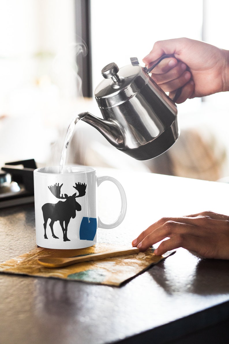 May include: A white ceramic mug with a black silhouette of a moose and a blue tea bag. A silver teapot is pouring hot water into the mug.