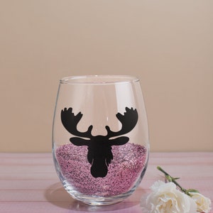 May include: A clear glass wine glass with a pink glitter base and a black moose silhouette decal.