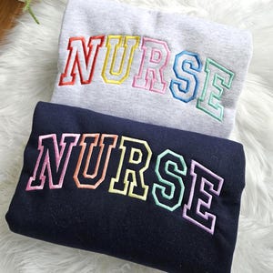 Puede incluir: Dos sudaderas, una gris y otra azul marino, bordadas con la palabra "NURSE" en letras coloridas de estilo universitario. La sudadera gris presenta bordados rojos, amarillos, rosas, azules y verdes. La sudadera azul marino tiene bordados rosas, naranjas, amarillos y verdes.