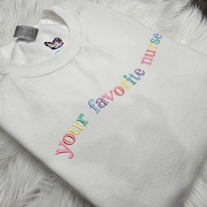 May include: White t-shirt with the text "Your favorite nurse" embroidered in rainbow colors.