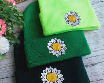 Puede incluir: Tres gorros de punto con margaritas blancas bordadas. El gorro de arriba es verde neón, el gorro del medio es verde oscuro y el gorro de abajo es negro.