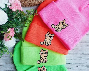 Puede incluir: Cuatro gorros de punto en diferentes colores, rosa, naranja, verde y verde lima, con un gato de dibujos animados bordado en cada uno.