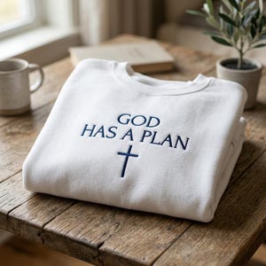 Peut inclure: Sweat-shirt blanc avec l'inscription brodée en bleu marine "GOD HAS A PLAN" et une croix bleue marine. Le sweat-shirt plié est posé sur une table en bois, avec une tasse, un livre et une petite plante en arrière-plan.