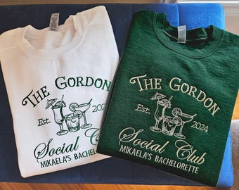 Może przedstawiać: Dwie białe i zielone bluzy z napisem "The Gordon Social Club" i rokiem "2024". Jedna bluza jest biała z zielonym napisem, a druga zielona z białym napisem. Obie bluzy mają grafikę koktajli z parasolkami.