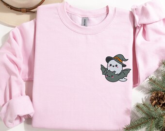 May include: A pink sweatshirt with a cute cartoon ghost wearing a witch hat and riding a bat. The ghost and bat are both smiling.