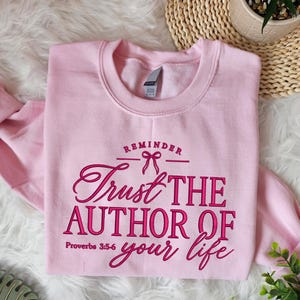 Peut inclure: Un sweat-shirt à col rond rose clair avec un texte rose foncé indiquant REMINDER, Trust THE AUTHOR OF your life, et Proverbs 3:5-6, accompagné d'un petit motif de nœud centré au-dessus du mot Trust. Le vêtement a des manches longues.