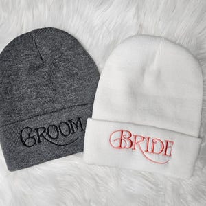 May include: Two knit beanies, one gray with the word "Groom" embroidered in black, and one white with the word "Bride" embroidered in pink.