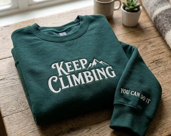 May include: A dark green sweatshirt with the words "KEEP CLIMBING" and a mountain graphic in white. The sleeve reads "YOU CAN DO IT". The sweatshirt is folded on a wooden surface, with a mug, plant, and blanket in the background.