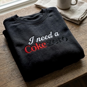 May include: Black sweatshirt featuring the embroidered text "I need a Coke Zero" in white and red. The garment is folded on a wooden surface, with a mug and folded cloth visible in the background.