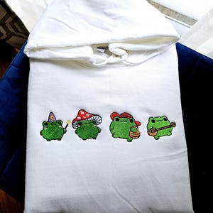Könnte beinhalten: Weißer Kapuzenpullover mit gestickten grünen Fröschen. Ein Frosch trägt einen Zaubererhut, ein anderer eine Pilzkappe, ein dritter einen Cowboyhut und der letzte spielt Ukulele. Die Frösche sind in Grün, Schwarz und Braun gestickt.