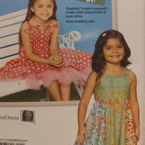 May include: Two young girls in dresses. One is wearing a red polka dot dress with a pink tulle skirt, and the other is wearing a green and floral patterned dress. The image includes the text "Simplicity create-it-yourself/crealo usted misma/créez-le vous-même www.simplicity.com."