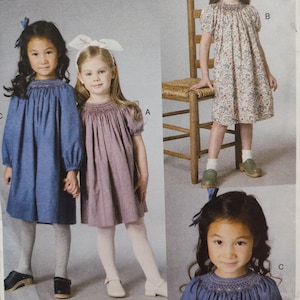 May include: Image of a sewing pattern featuring three little girls in dresses. Two girls are standing side-by-side, wearing dresses in blue and mauve. The third girl is standing next to a wooden chair, wearing a floral print dress. All dresses have embroidered necklines.