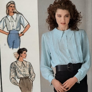 May include: A light blue long-sleeve button-down shirt with a pointed collar and vertical pleats along the front placket. The shirt is paired with a wide black belt featuring a large circular buckle worn over a black skirt or pair of trousers.