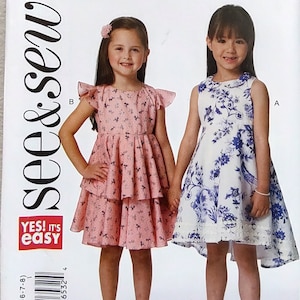 May include: A sewing pattern for girls' dresses. The pattern features two dress designs: one with a tiered skirt and a pink floral print, and another with a blue and white floral print. The text "see & sew" and "YES! IT'S easy" are visible.