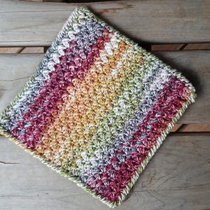 May include: A square, hand-crocheted washcloth with a striped pattern in shades of red, yellow, green, and white. The edges have a green border. The washcloth is on a weathered wooden surface.