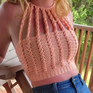 May include: Peach-colored crochet halter top with a unique openwork design. The top features vertical braided straps and a textured, fitted bodice. The handmade garment is a crop top style, perfect for warm weather. The top is paired with blue denim jeans.