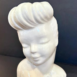 May include: A glossy white ceramic head planter featuring a stylized figure with closed eyes and a prominent, swept-back hairstyle. The sculpture depicts the head and shoulders of a person with smooth, reflective surfaces and a small floral detail.