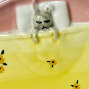 Puede incluir: Un plato de cer&aacute;mica con un dise&ntilde;o de conejo durmiendo. El conejo es gris y descansa sobre una almohada blanca bajo una manta amarilla con detalles florales amarillos. El borde del plato es rosa.