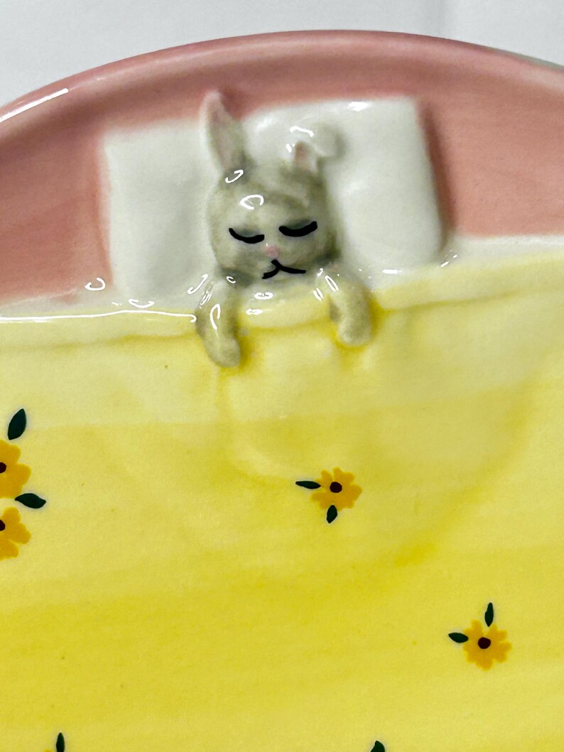 Puede incluir: Un plato de cer&aacute;mica con un dise&ntilde;o de conejo durmiendo. El conejo es gris y descansa sobre una almohada blanca bajo una manta amarilla con detalles florales amarillos. El borde del plato es rosa.