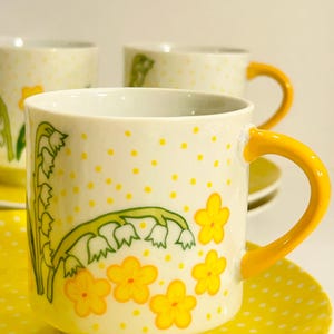 May include: A white ceramic mug with a yellow handle, featuring a pattern of small yellow polka dots, green lily of the valley stems, and yellow five-petaled flowers. The mug sits on a matching saucer with yellow polka dots on a white background.