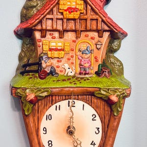 May include: A wall clock featuring a three-dimensional cottage scene with a red roof, brick walls, and two figures with a dog. The clock face is circular with black numerals and ornate gold hands, framed by a wood-textured base with acorn accents.