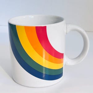 May include: A white ceramic mug featuring a curved, multi-colored rainbow graphic on the front. The design consists of four distinct, thick stripes in shades of dark blue, dark green, yellow, orange, and red, arranged in a sweeping arc pattern.