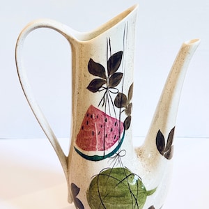 May include: A tall, cream-colored ceramic pitcher with a speckled finish, featuring a long handle and a slender spout. The body is decorated with a hand-painted illustration of a pink watermelon slice and a green melon, accented by dark brown leaves.