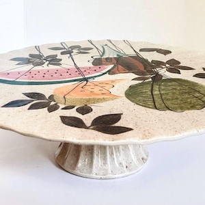 May include: A round ceramic cake stand with a scalloped edge and a pedestal base. The surface features a speckled cream finish decorated with painted illustrations of a watermelon slice, a cantaloupe wedge, a green squash, and dark leaves.