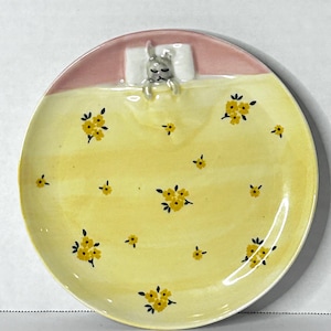 Puede incluir: Un plato de cer&aacute;mica con un dise&ntilde;o caprichoso. La parte superior es rosa, con una almohada blanca y un conejito durmiendo. La parte inferior es amarilla, decorada con patrones florales amarillos. Una pieza decorativa &uacute;nica.