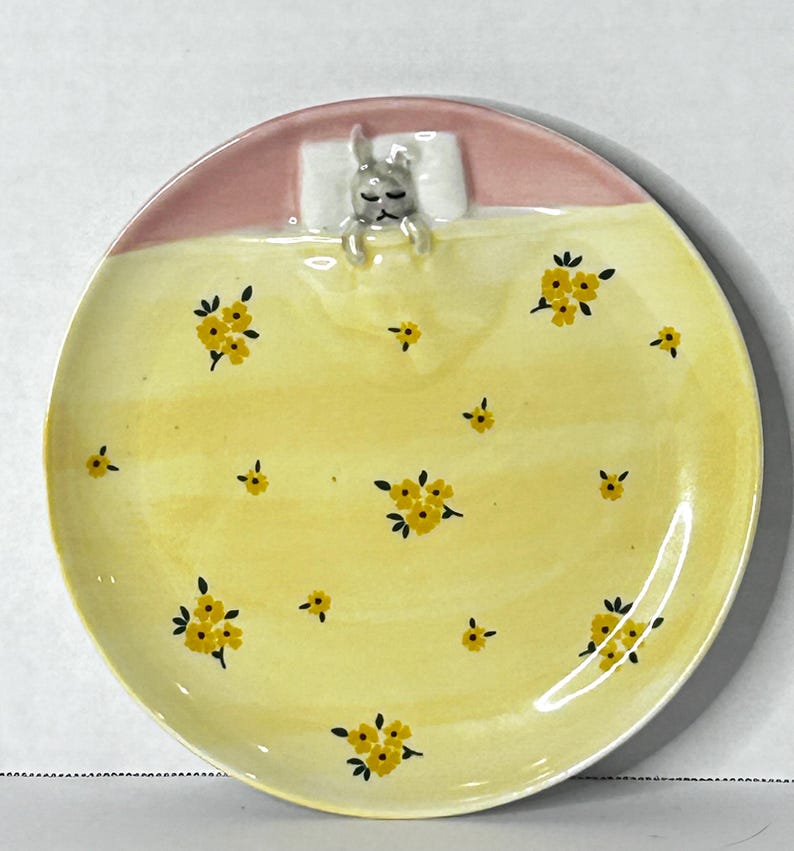 Puede incluir: Un plato de cer&aacute;mica con un dise&ntilde;o caprichoso. La parte superior es rosa, con una almohada blanca y un conejito durmiendo. La parte inferior es amarilla, decorada con patrones florales amarillos. Una pieza decorativa &uacute;nica.