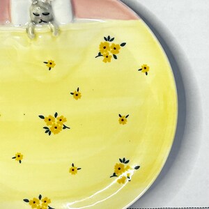Puede incluir: Plato de cer&aacute;mica con dise&ntilde;o de conejito durmiente. El plato presenta una superficie amarilla con motivos florales amarillos y un borde rosa. El conejito descansa sobre una almohada blanca bajo una manta amarilla.