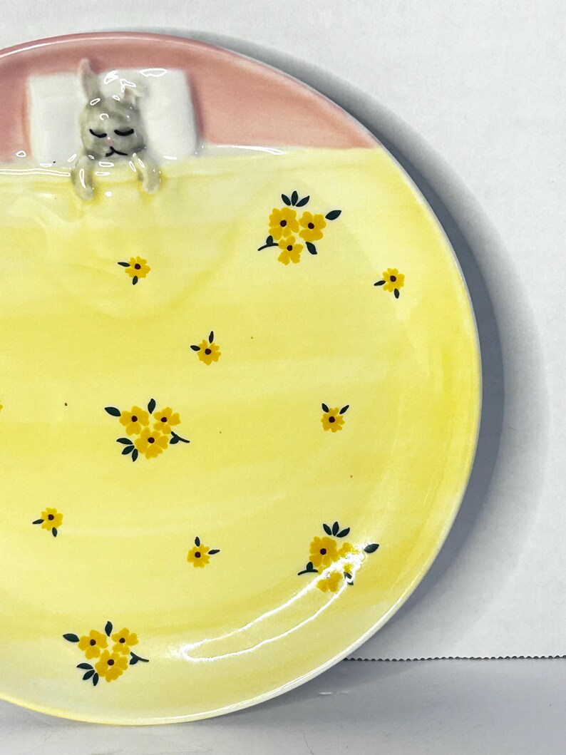 Puede incluir: Plato de cer&aacute;mica con dise&ntilde;o de conejito durmiente. El plato presenta una superficie amarilla con motivos florales amarillos y un borde rosa. El conejito descansa sobre una almohada blanca bajo una manta amarilla.