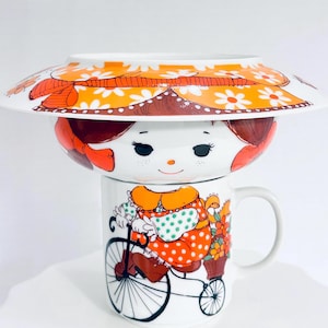 May include: A white ceramic mug and bowl set featuring a graphic illustration of a character with brown hair wearing an orange hat with white daisies. The mug depicts the same character riding a bicycle with a basket of orange flowers.