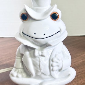 May include: A white ceramic frog lamp shaped like a gentleman wearing a top hat, a tuxedo jacket, and a bow tie. The frog has large, round, orange-rimmed eyes with black pupils and a thin black line for a mouth. A white power cord extends from the side.