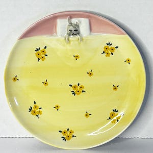 Puede incluir: Plato de cer&aacute;mica decorativo con un dise&ntilde;o caprichoso. El plato presenta una superficie amarilla adornada con patrones florales amarillos. Un conejito durmiente est&aacute; acurrucado en una almohada blanca en la parte superior, con un borde rosa. Ideal para servir aperitivos o como pieza decorativa.