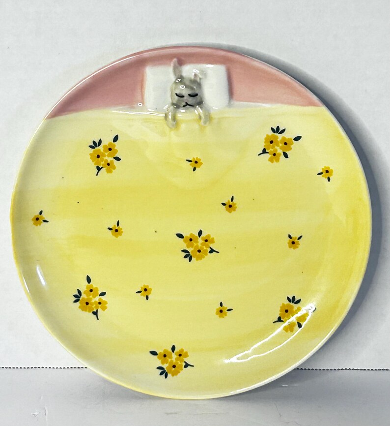 Puede incluir: Plato de cer&aacute;mica decorativo con un dise&ntilde;o caprichoso. El plato presenta una superficie amarilla adornada con patrones florales amarillos. Un conejito durmiente est&aacute; acurrucado en una almohada blanca en la parte superior, con un borde rosa. Ideal para servir aperitivos o como pieza decorativa.