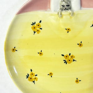 Puede incluir: Un plato de cer&aacute;mica con un dise&ntilde;o caprichoso. El plato presenta una figura de conejito durmiendo bajo una manta rosa y una almohada blanca. La superficie amarilla est&aacute; decorada con patrones florales amarillos. Una pieza encantadora y decorativa.