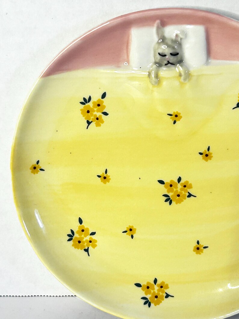 Puede incluir: Un plato de cer&aacute;mica con un dise&ntilde;o caprichoso. El plato presenta una figura de conejito durmiendo bajo una manta rosa y una almohada blanca. La superficie amarilla est&aacute; decorada con patrones florales amarillos. Una pieza encantadora y decorativa.