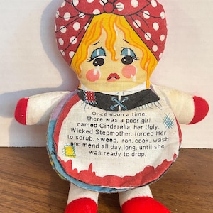 May include: A handmade Cinderella doll with a red and white polka dot headscarf and a sad expression. The doll has a white apron with text describing Cinderella's story. The doll's arms and legs are red.