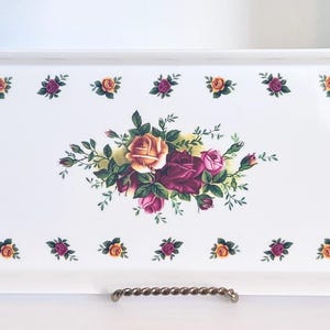 May include: A rectangular white ceramic serving tray featuring a central floral arrangement of yellow, pink, and deep red roses with green leaves. The border is decorated with a repeating pattern of small rose clusters in matching colors.