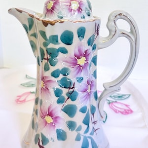 May include: A porcelain coffee pot featuring a white base with hand-painted pink flowers, yellow centers, and teal green leaves. The pot has a scalloped rim, a curved handle, a matching lid with a finial, and gold-toned trim along the edges.
