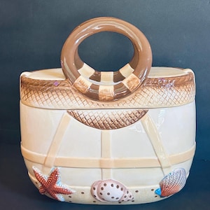 May include: A ceramic cookie jar shaped like a beach tote bag with a glossy cream finish. The bag features a brown circular handle, a woven-texture top rim, and raised decorative elements including a red starfish and two seashells.