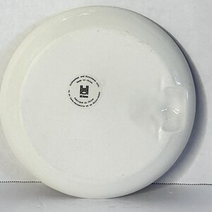 Puede incluir: Un plato de cer&aacute;mica blanco con un borde ligeramente elevado. La parte inferior presenta un sello circular con texto, incluyendo "Apto para lavavajillas y microondas" y "Hecho en China". El plato tiene un acabado liso y brillante.