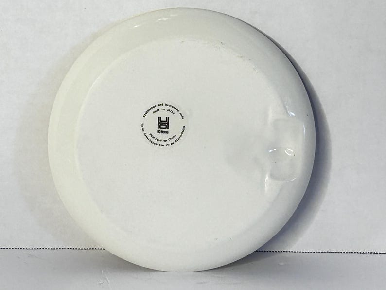 Puede incluir: Un plato de cer&aacute;mica blanco con un borde ligeramente elevado. La parte inferior presenta un sello circular con texto, incluyendo "Apto para lavavajillas y microondas" y "Hecho en China". El plato tiene un acabado liso y brillante.