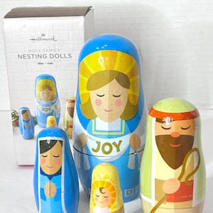 May include: A set of Hallmark Holy Family nesting dolls. The dolls are painted with bright colors and feature the figures of Mary, Joseph, and baby Jesus. The largest doll is blue with the word "JOY" on it.